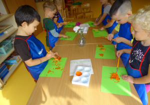 Dzieci stoją przy stoliku i malują liście pomarańczową farbą